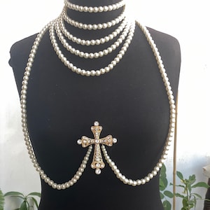 Puede incluir: Un collar de perlas blancas con un colgante de cruz dorada adornado con diamantes de imitación transparentes. El collar está en capas y drapeado sobre un maniquí negro.