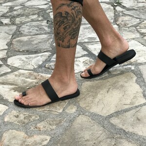 May include: A pair of black leather thong sandals with a single strap across the foot. The sandals are worn on a person's feet, and the person has a tattoo on their lower leg.
