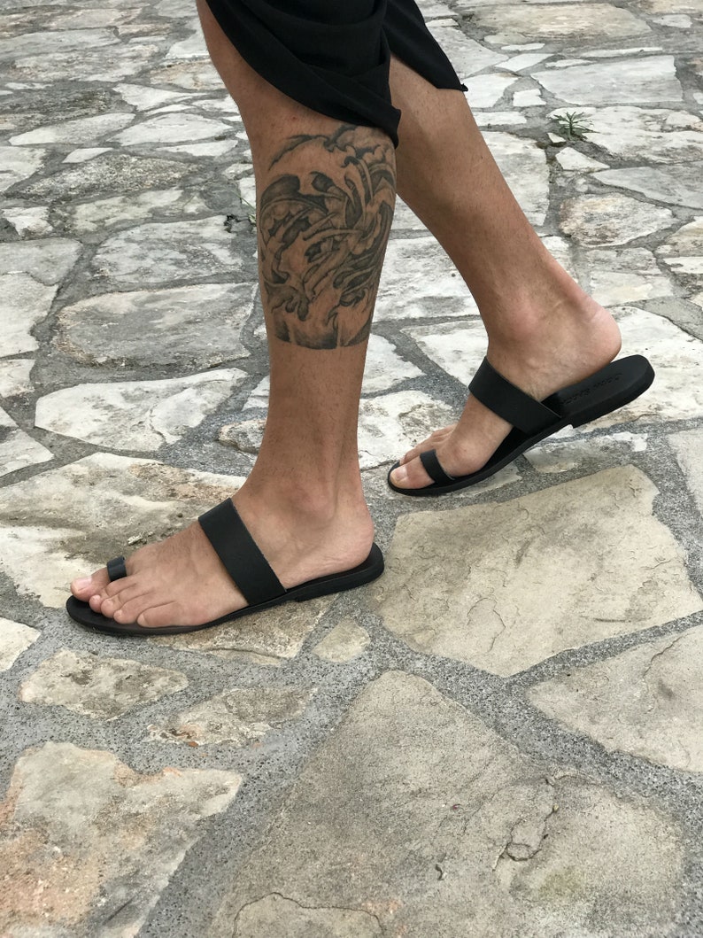 May include: A pair of black leather thong sandals with a single strap across the foot. The sandals are worn on a person's feet, and the person has a tattoo on their lower leg.
