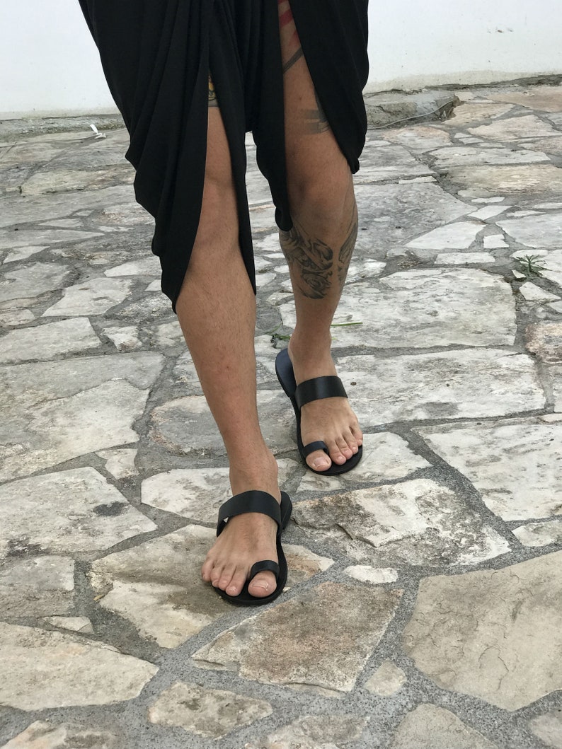 May include: A pair of black leather sandals with a toe ring and a strap across the foot. The sandals are worn on a person's feet, standing on a stone surface.