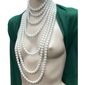 May include: A collection of white faux pearl necklaces of varying lengths, displayed on a mannequin. The necklaces create a layered effect. The mannequin is set against a green fabric background, which highlights the white pearls. The pearls are round and evenly sized.