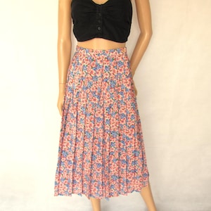 May include: A black crop top with a sweetheart neckline and button details paired with a floral print pleated midi skirt. The skirt features a pattern of pink, blue, and white flowers. The model is wearing black sandals.