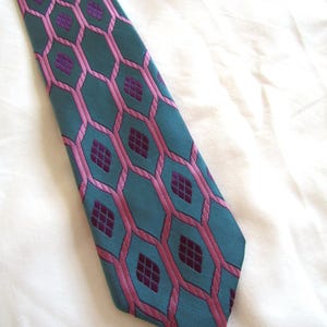 Puede incluir: Una corbata con fondo verde azulado y un patrón repetitivo de contornos hexagonales rosas. Dentro de cada hexágono rosa hay una forma de diamante de color púrpura oscuro que contiene un pequeño patrón de cuadrícula. La corbata tiene un extremo puntiagudo.