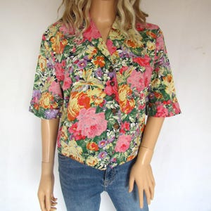 Peut inclure: Une chemise à fleurs à manches courtes avec un motif vibrant de fleurs roses, jaunes, oranges et violettes sur un fond vert. La chemise a un col classique et des boutons en bois. Le mannequin porte un jean bleu.