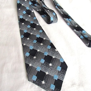 Puede incluir: Corbata estampada con un diseño repetitivo de cuadrados negros, cuadrados azul claro y detalles blancos sobre un fondo gris. La corbata está hecha de tela lisa, adecuada para vestimenta formal o de negocios.