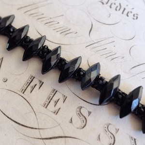 Op de afbeelding: Een close-up van een zwarte kralenarmband met facetgeslepen, marquise-vormige kralen. De armband ligt op een vintage papieren achtergrond met elegant schrift en het woord "FESS" zichtbaar.
