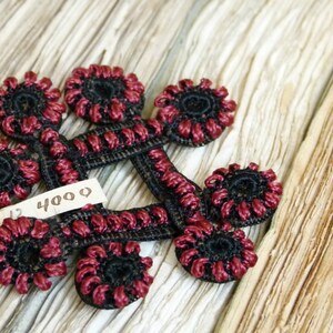 Puede incluir: Primer plano de una colección de adornos florales negros y burdeos. Los adornos están dispuestos en un patrón circular y están hechos de un material tejido. Una pequeña etiqueta con el número "4000" es visible. El fondo es una superficie clara y texturizada.