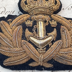 Puede incluir: Un parche bordado azul marino vintage con un diseño de ancla dorada, corona y corona de laurel. Los intrincados detalles incluyen hilo dorado y un reverso de fieltro negro. Este parche es probablemente un emblema militar o náutico.