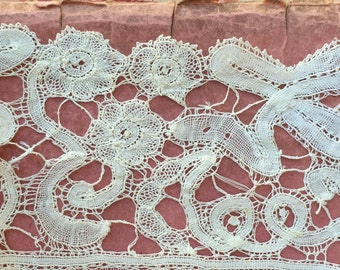 May include: Close-up of cream-colored lace with floral and scroll designs. The lace features detailed openwork patterns, including flowers and swirling motifs. The delicate fabric is set against a pink background, highlighting the intricate craftsmanship.