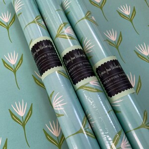 May include: Three rolls of turquoise blue wrapping paper with a white floral pattern. The paper is labeled "Rebecca Jane Wellbeing" and "Wrapping Paper".