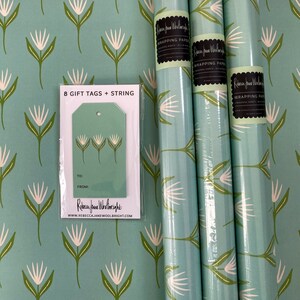 May include: Three rolls of teal blue wrapping paper with a white floral pattern. The paper is packaged with 8 gift tags and string. The tags are teal blue with a white floral design and say "To:" and "From:" on them. The text on the wrapping paper says "Rebecca Jane Woolbright Wrapping Paper".