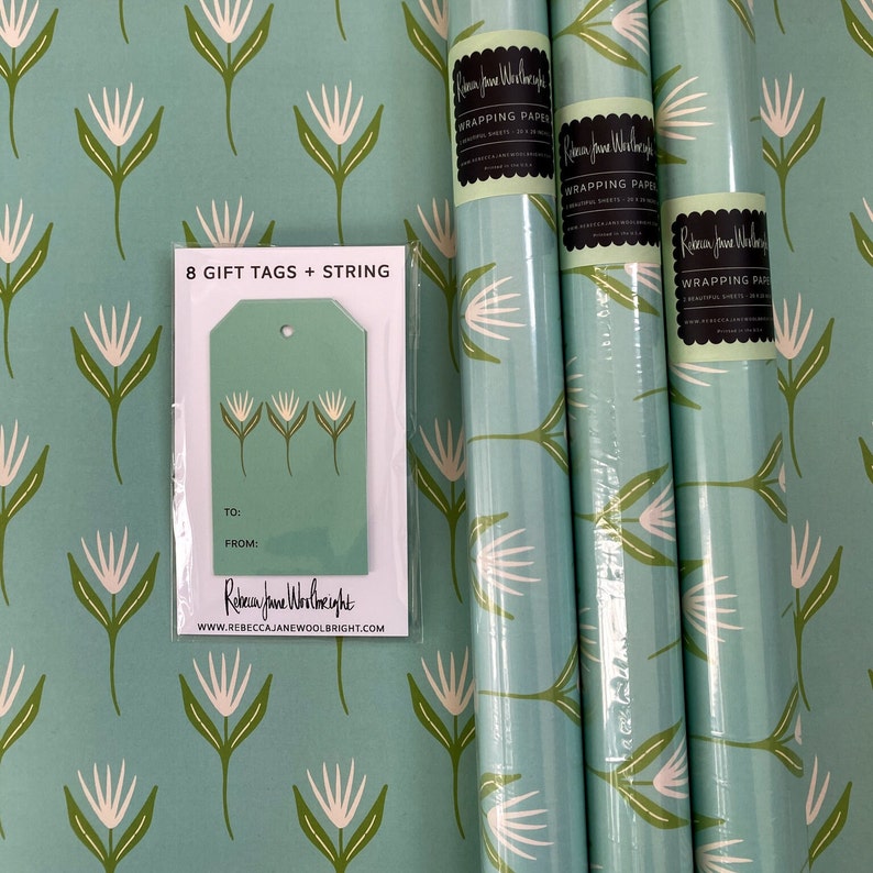 May include: Three rolls of teal blue wrapping paper with a white floral pattern. The paper is packaged with 8 gift tags and string. The tags are teal blue with a white floral design and say "To:" and "From:" on them. The text on the wrapping paper says "Rebecca Jane Woolbright Wrapping Paper".