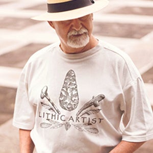 May include: A man wearing a white t-shirt with a graphic of a mushroom and the text "Lithic Artist". He is also wearing a straw hat with a black band.