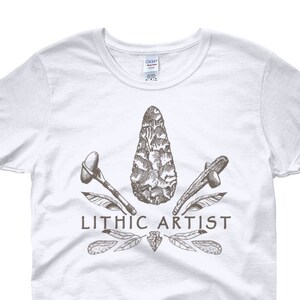 May include: White women's t-shirt with a brown graphic of a stone tool, feathers, and the text "Lithic Artist".