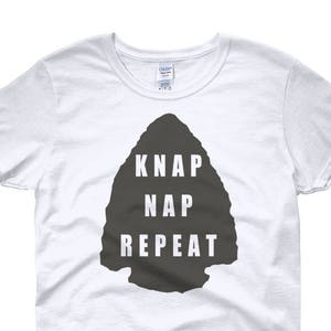 May include: White women's t-shirt featuring a grey arrowhead graphic with the text "KNAP NAP REPEAT".