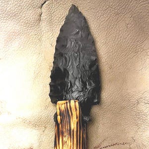 Puede incluir: Una punta de flecha de obsidiana negra con un mango de madera. La punta de flecha es puntiaguda y tiene una superficie lisa y pulida. El mango es de madera y tiene un acabado natural y rústico.