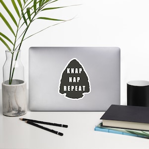 May include: A black and white sticker with a grey arrowhead shape. The arrowhead has the words "KNAP NAP REPEAT" written inside.