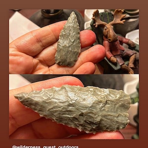 May include: Two gray stone arrowheads, one triangular and one elongated, are held in a hand. The arrowheads have a rough, textured surface.