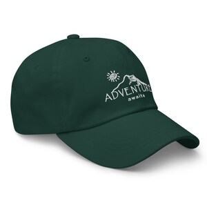 Puede incluir: Una gorra de b&eacute;isbol verde oscuro con un dise&ntilde;o bordado en blanco de una monta&ntilde;a, un sol y el texto "ADVENTURE awaits".