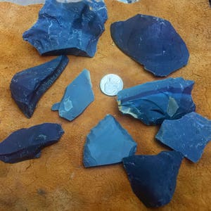 May include: A collection of dark blue and gray obsidian flakes and fragments, with a silver coin for scale, arranged on a brown leather surface. The obsidian pieces vary in size and shape, showcasing natural textures and edges.