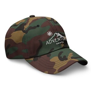 Puede incluir: Una gorra de b&eacute;isbol de camuflaje verde, marr&oacute;n y negro con un dise&ntilde;o bordado en blanco que dice "ADVENTURE awaits" con un sol y una monta&ntilde;a.