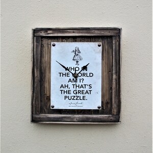 May include: A wooden framed clock with a white face and black hands. The clock face features a black and white illustration of Alice from Alice in Wonderland and the text "Who in the world am I? Ah, that's the great puzzle."  The text is attributed to Lewis Carroll.