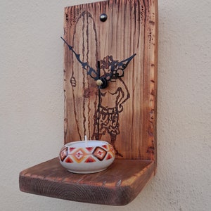 May include: Wooden wall clock with a shelf underneath. The clock face features a surfer silhouette and the hands are black. A small candle holder with a white base and orange and red geometric pattern is on the shelf.