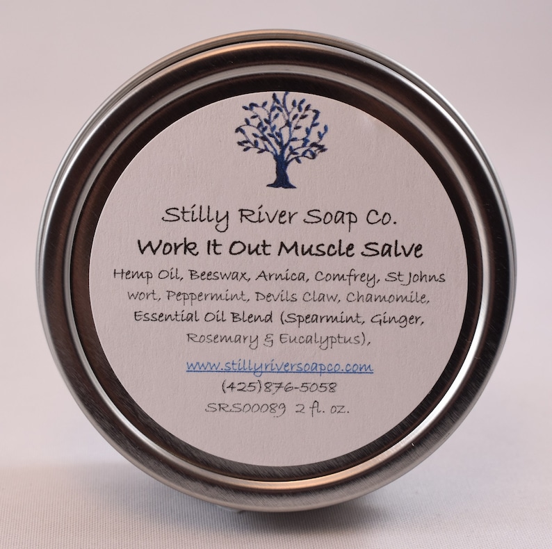 May include: A silver tin with a white label that reads "Stilly River Soap Co. Work It Out Muscle Salve" and lists the ingredients: Hemp Oil, Beeswax, Arnica, Comfrey, St Johns wort, Peppermint, Devils Claw, Chamomile, Essential Oil Blend (Spearmint, Ginger, Rosemary & Eucalyptus). The label also includes the company website, phone number, and product code: SRS00089. The tin contains 2 fluid ounces of salve.