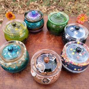 May include: A collection of seven decorative glass jars with lids, each featuring unique colour schemes and designs. The jars are arranged on a wooden surface, with floral accents. The jars have a diameter of approximately 7.6 cm.