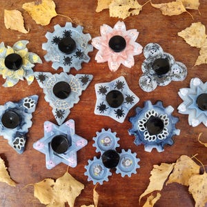 May include: A collection of decorative resin fidget spinners in various shapes, including gears, flowers, and stars. Each spinner features a black center and is decorated with spiders, paw prints, and other designs. The spinners are in shades of blue, pink, and gray.