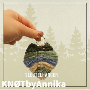 May include: A macrame keychain in shades of green, blue, and tan, held by a hand. The keychain has a leaf-like design and is attached to a silver keyring. The text "SLEUTELHANGER" and "KNØTbyAnnika" are visible.