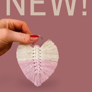 May include: A cream and pink heart-shaped macrame keyring, held against a mauve background. The word "NEW!" is displayed in large, light-coloured letters above the keyring. The keyring is made of woven cotton.