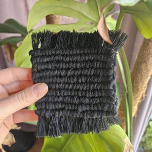 May include: A square, black macrame coaster with a fringed edge. The coaster is made of woven black cotton cord. The background includes green Monstera leaves and a brown plant support.