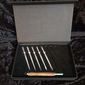 May include: A set of crochet hooks and tools presented in a gray box with a black interior. The hooks are silver with wooden handles. The set includes various hook sizes and a wooden-handled tool.