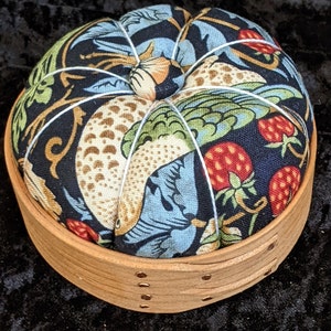 May include: A round, fabric-covered pincushion with a floral and bird design in shades of blue, green, brown, and red. The pincushion is nestled in a wooden base with a natural finish.