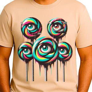 May include: A tan t-shirt featuring a graphic of five colorful lollipops with eye designs. The lollipops have a swirl of colors including pink, blue, yellow, and green. The person in the image is smiling.