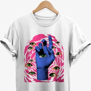 Puede incluir: Camiseta blanca con un diseño de mano azul, un ojo en la palma y múltiples ilustraciones con ojos verdes. El diseño está sobre un fondo arremolinado rosa y blanco.