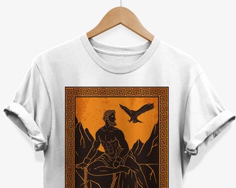 May include: White t-shirt with a graphic print of a man chained to a rock with an eagle flying above him. The graphic is in a brown and orange color scheme and is framed with a Greek key border.
