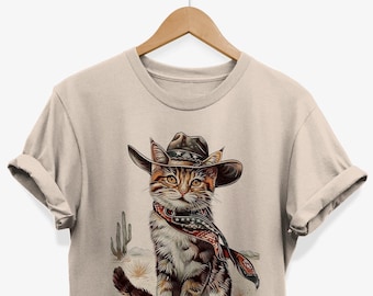 May include: A beige t-shirt with a graphic of a cat wearing a cowboy hat and bandana. The cat is sitting in a desert scene with cacti.