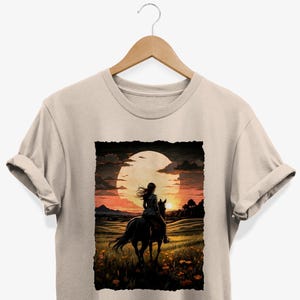 Könnte beinhalten: Ein beiges T-Shirt mit einem grafischen Aufdruck einer Person, die auf einem Pferd in den Sonnenuntergang reitet. Die Sonne ist ein großer orangefarbener Kreis am Himmel.