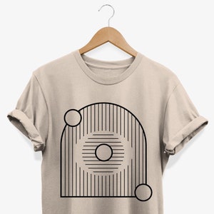 May include: A beige t-shirt with a black geometric design featuring a circle within a striped arch and two smaller circles.
