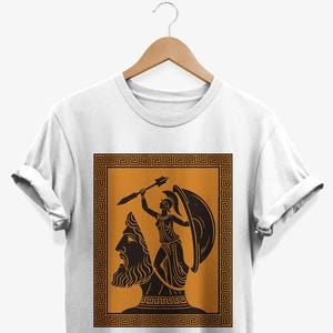 May include: White t-shirt with a black and orange graphic depicting a classical Greek scene. The design shows a bearded bust and a figure with a spear and shield, within a decorative border. The t-shirt is displayed on a wooden hanger.