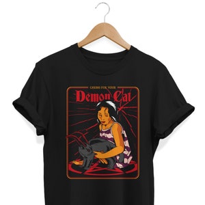 May include: Black t-shirt with a graphic of a girl holding a black cat in a pentagram. The graphic is in a red frame with the text "Caring for your Demon Cat".