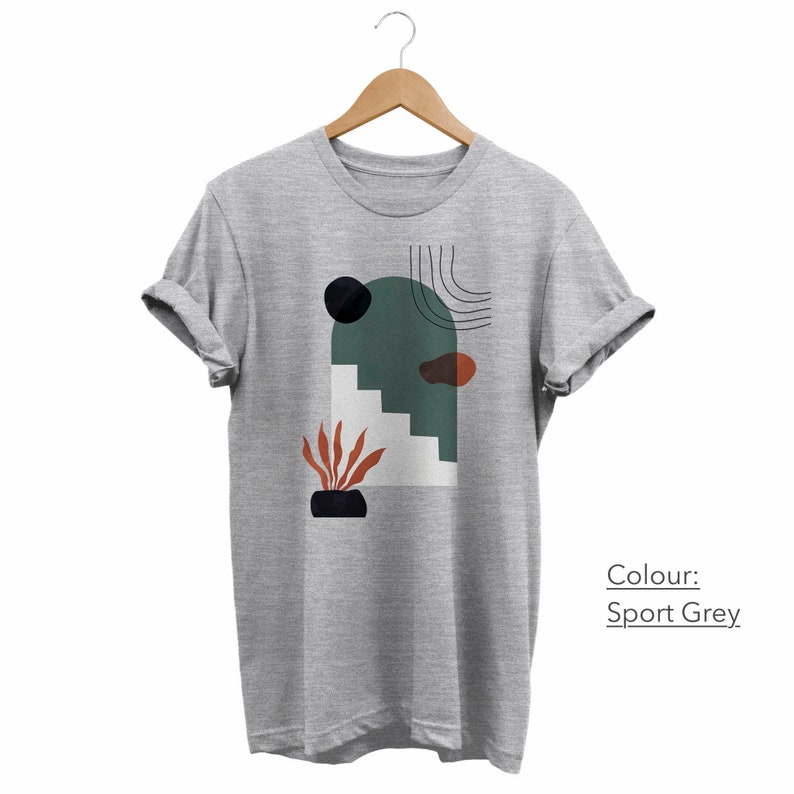May include: A gray t-shirt with a graphic print of an abstract design featuring a green arch, stairs, a black circle, and a red and orange plant. The text "Colour: Sport Grey" is visible below the graphic.
