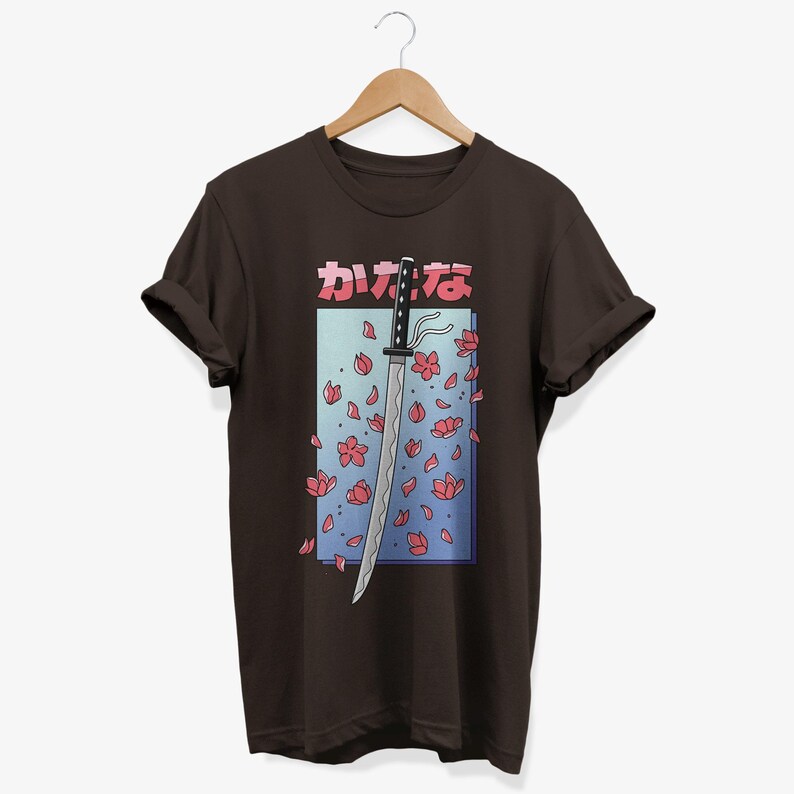 May include: A brown t-shirt featuring a graphic of a katana sword with pink cherry blossoms falling around it. The Japanese text on the shirt reads "かなな".