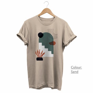 May include: A sand colored t-shirt with a graphic print of an abstract design featuring a green arch, stairs, a black circle, a red and orange shape, and a plant in a black pot. The text "Colour: Sand" is printed below the graphic.