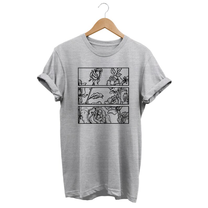 May include: A light gray t-shirt with a black floral design. The design features three rectangular panels, each showcasing a different section of a rose illustration. The shirt is hanging on a wooden hanger.