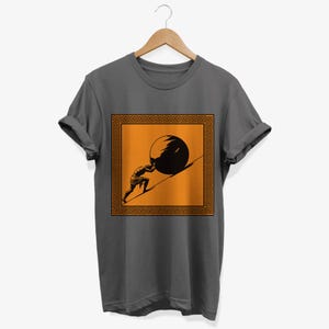 May include: A dark gray t-shirt with a graphic of a person pushing a large stone uphill. The graphic is framed by a square border with a repeating Greek key pattern in orange. The background of the graphic is orange.