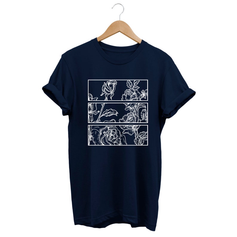 May include: Navy blue t-shirt with a white floral design. The design is divided into three rectangular sections, each showcasing detailed white line art of roses and leaves. The shirt is displayed on a wooden hanger.
