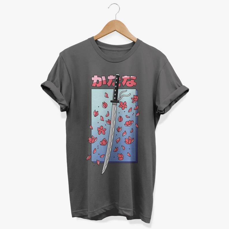 May include: A dark grey t-shirt featuring a graphic of a katana sword with pink cherry blossoms falling around it. The Japanese text "カワイイ" is printed above the sword.